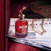 Red perfume bottle labeled 'Scandal' on a newspaper with a gold necklace beside it.